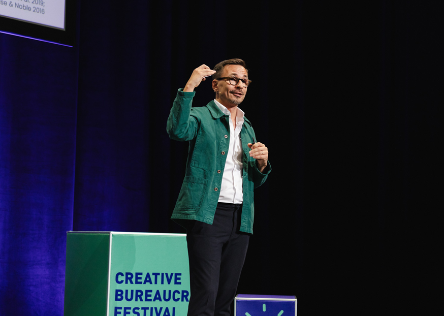 Tommi Laitio wearing a green jacket stands on stage and raises one hand