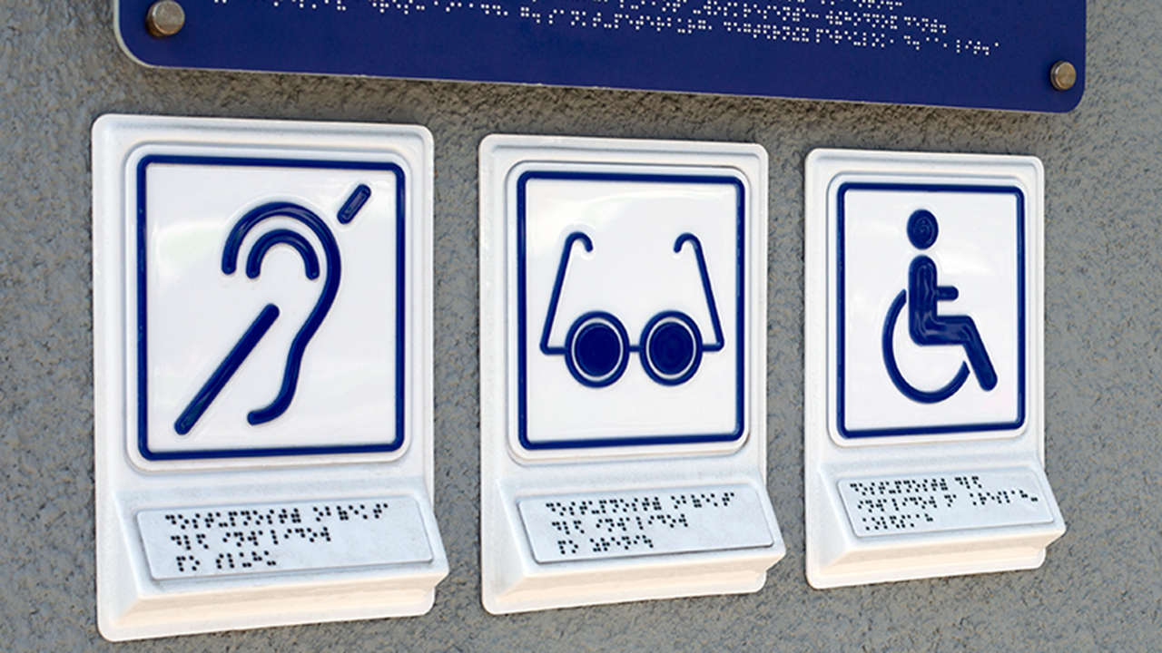 Five Addressable Accessibility Gaps In Cities Accessibility signage in an urban environment, including the international symbols of access for hearing loss, access for low vision, and the wheelchair symbol for individuals with limited mobility.